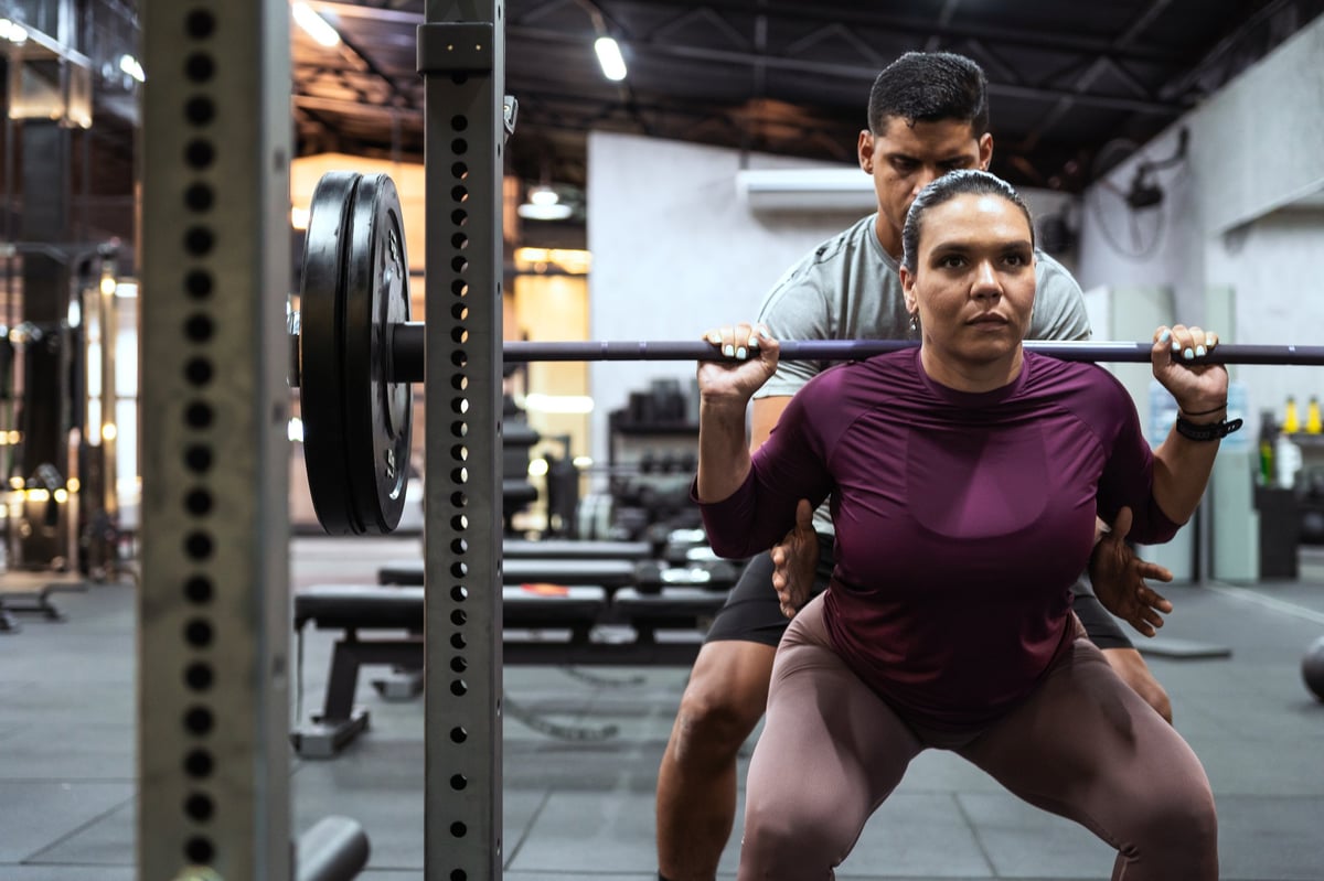 Latin american woman performing barbell squats with the assistance of a male personal trainer at a fitness gym